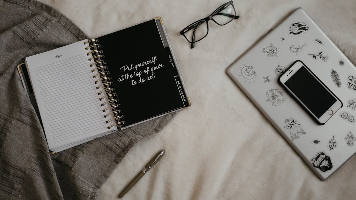 A cozy flat lay featuring a notebook, smartphone, laptop with designs, and eyeglasses on bedspread.